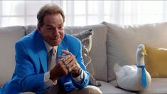 a man in a blue suit sitting on a couch next to two white ducks and one is wearing a ring