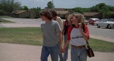 two young people holding hands walking down the sidewalk in front of some parked cars and houses