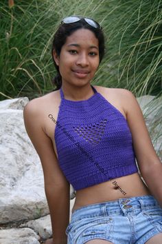 a young woman sitting on top of a rock next to tall grass and rocks wearing a purple crocheted halter top