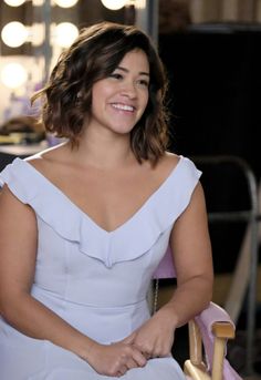 a smiling woman in a white dress sitting on a pink chair and looking at the camera