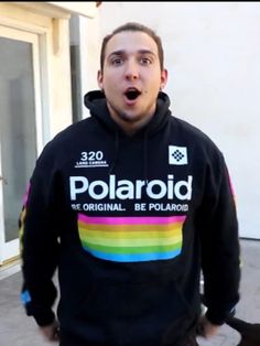 a man in a black polaroid hoodie is making a surprised face while standing outside