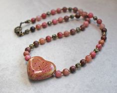 Pink and bronze heart necklace is made with a unique, pink and brown ceramic heart bead on a necklace of natural pink with black inclusions rhodonite stones separated by bronze seed beads. Bronze color lobster clasp. ❖  30x25mm ceramic heart bead ❖  6mm rhodonite stones ❖  4mm bronze and crystal hexagon beads ❖  glass seed beads ❖  bronze color lobster clasp   Necklace is 18" in length. Heart-shaped Beaded Necklaces With Natural Stones, Heart-shaped Beaded Necklaces With Natural Stones For Gift, Heart-shaped Natural Stone Beaded Necklace For Gift, Heart-shaped Beaded Necklace With Natural Stones For Gift, Pink Heart Pendant Beaded Necklace As Gift, Pink Heart Pendant Beaded Necklace For Gift, Pink Beaded Necklace With Heart Pendant As Gift, Pink Heart Pendant Beaded Necklace For Valentine's Day, Pink Heart-shaped Bohemian Beaded Necklaces