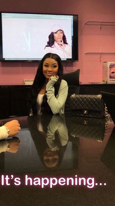 a woman sitting at a table in front of a large screen with the words it's happening