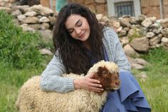 a woman holding a lamb in her lap while sitting on the ground outside with rocks and grass behind her