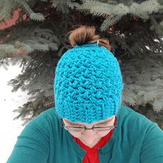 a woman wearing a blue crocheted beanie and reading a book in front of a pine tree