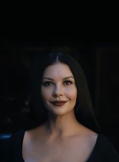 a woman with long black hair wearing a black dress and smiling at the camera while standing in front of a dark background