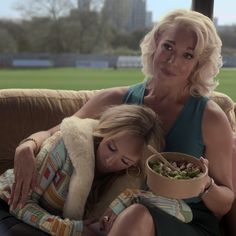 two women are sitting on a couch and one is holding a bowl with food in it