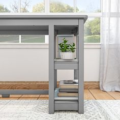 a grey desk with a plant on it in front of a window and white curtains