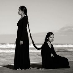 two women are sitting on the beach and one is holding her long hair in braids