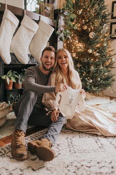 a man and woman sitting on the floor in front of a christmas tree with stockings