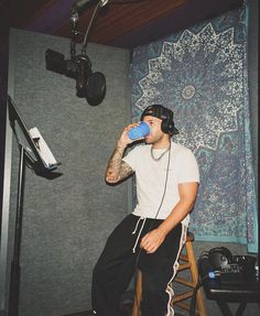 a man sitting on a stool drinking from a blue cup while listening to headphones