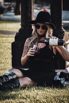 a woman sitting on the ground with a glass of wine in her hand and wearing a hat