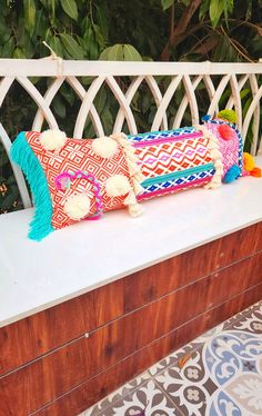 three colorful pillows sitting on top of a wooden bench next to a wall and trees