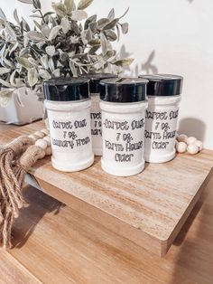 three white jars with black labels on them sitting on a wooden table next to a potted plant