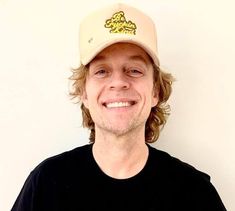 a man with long hair wearing a black shirt and a baseball cap smiles at the camera