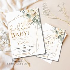 two baby shower cards sitting on top of a white bed next to flowers and greenery