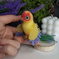 a hand holding a small yellow bird made out of beaded beads with purple flowers in the background