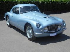 an old blue sports car parked in a parking lot next to some hedges and bushes