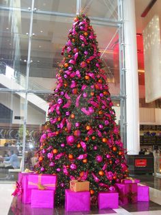 a large christmas tree with pink presents in front of it and people walking around the building