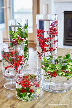 three glass vases filled with red berries and greenery on a table in front of a fireplace