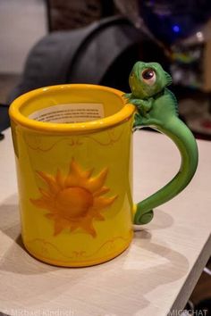 a yellow coffee mug with a green lizard on it's side sitting on a table