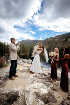 a bride and groom kiss on the rocks by the water as their wedding party looks on