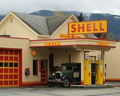 an old car is parked in front of a shell gas station with mountains in the background