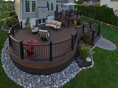 a deck with chairs and tables in front of a house surrounded by rocks, grass and flowers