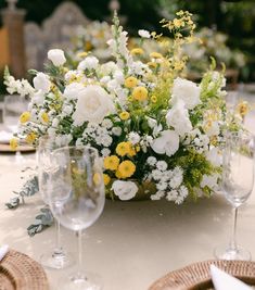 an arrangement of white and yellow flowers in a basket on a table with wine glasses