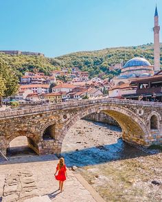 a woman in a red dress is standing on a stone bridge over a small river