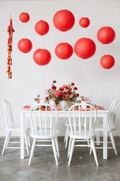 a table with white chairs and red lanterns hanging from the wall above it, surrounded by flowers