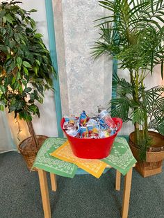 a red bucket filled with candy sitting on top of a table next to potted plants