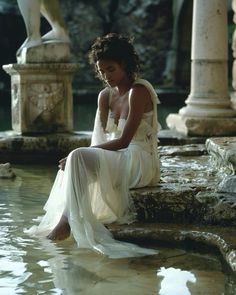 a woman sitting on the edge of a body of water wearing a white dress and sandals