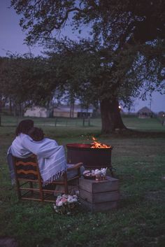 two people sitting in chairs around an open fire pit at night with the lights on