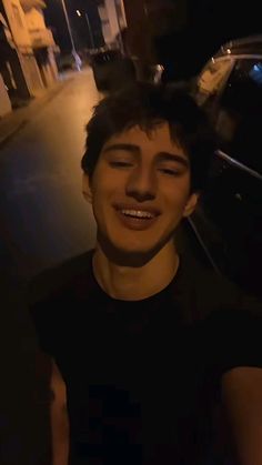 a young man smiling while standing next to a parked car on a street at night