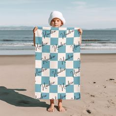 a little boy holding up a blue and white checkered blanket on the beach with his name written in cursive writing