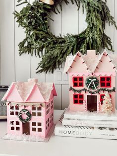 two pink christmas houses sitting on top of books next to a wreath and pine tree