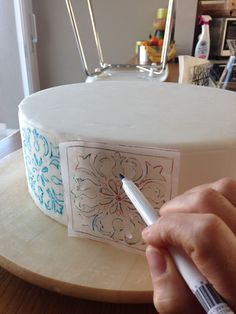 a person is writing on a cake with a marker in their left hand and the other hand holding a pen