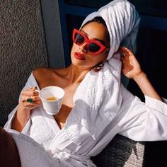 a woman wearing red sunglasses and a white robe holding a cup of coffee while sitting on a chair