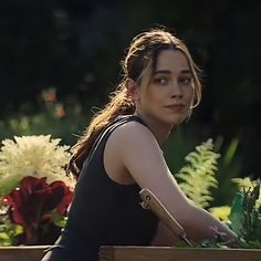 a woman sitting on a bench with flowers in the back ground and behind her is a wooden box