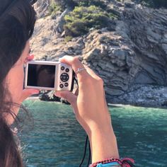 a woman taking a photo with her cell phone near the water's edge and cliffs in the background