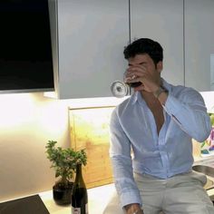 a man drinking from a wine glass while sitting on top of a kitchen counter next to a potted plant