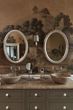 two sinks with mirrors above them in front of a wallpapered mural behind them