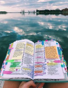 someone is holding an open bible in their hand while sitting on a dock by the water