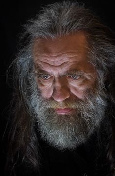 an old man with long hair and beard looking at the camera, in front of a black background