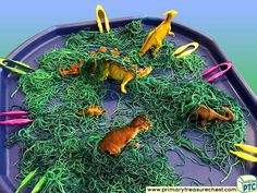 some plastic dinosaurs are in the grass on a blue tray with yellow and pink scissors