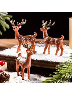 three small deer figurines sitting on top of a snow covered table next to pine cones