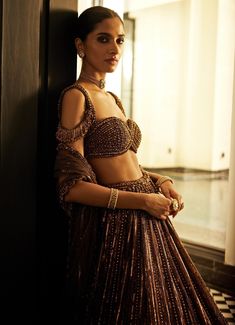 Elevate your wardrobe with this breathtaking Copper Crystal Linear Embroidered Organza Lehenga Set, a masterpiece of intricate craftsmanship perfect for making a bold style statement. Made from luxurious organza fabric, the copper lehenga features exquisite linear embroidery adorned with sequins, cutdana, and crystal embellishments, adding a radiant sparkle to the flowing design. Paired with a jewelled georgette blouse, the lehenga set offers a modern twist with its chic cut and crystal embroidery. The rich copper hue is beautifully complemented by the matching tulle dupatta, intricately detailed with shimmering accents. Ideal for weddings, receptions, or cocktail. The off shoulder tassels details can be added for an additional GBP 45. Composition : Lehenga - Organza, Dupatta - Soft Net an Straight Lehenga, Copper Lehenga, Linear Embroidery, Shoulder Tassels, Vani Vats, Organza Lehenga, Crystal Embroidery, Weddings Receptions, Copper Crystal