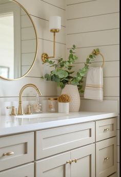 a bathroom with white cabinets and gold accessories on the counter top, along with a round mirror