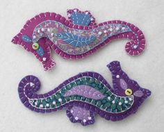 two purple and green handmade items on a white table top, one is shaped like a lizard